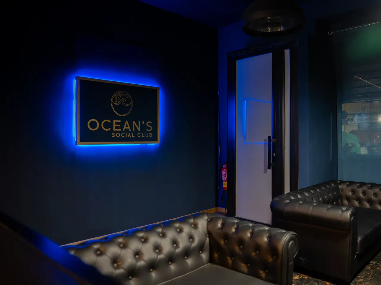 The OCEAN’S SOCIAL CLUB sign, backlit in blue on a navy wall, above black chesterfield sofas
